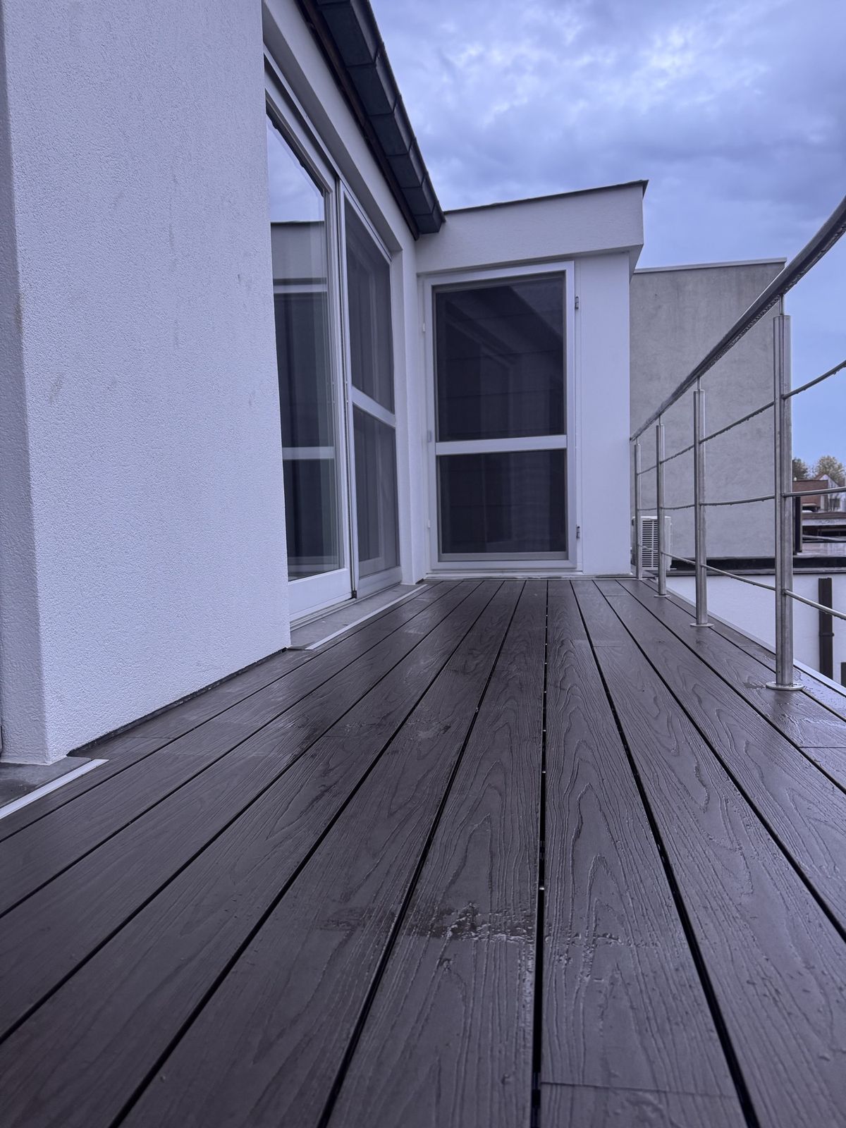 Het nieuwe terras in donkere planken langs een witte gevel, met een inox reling en een hoekzone